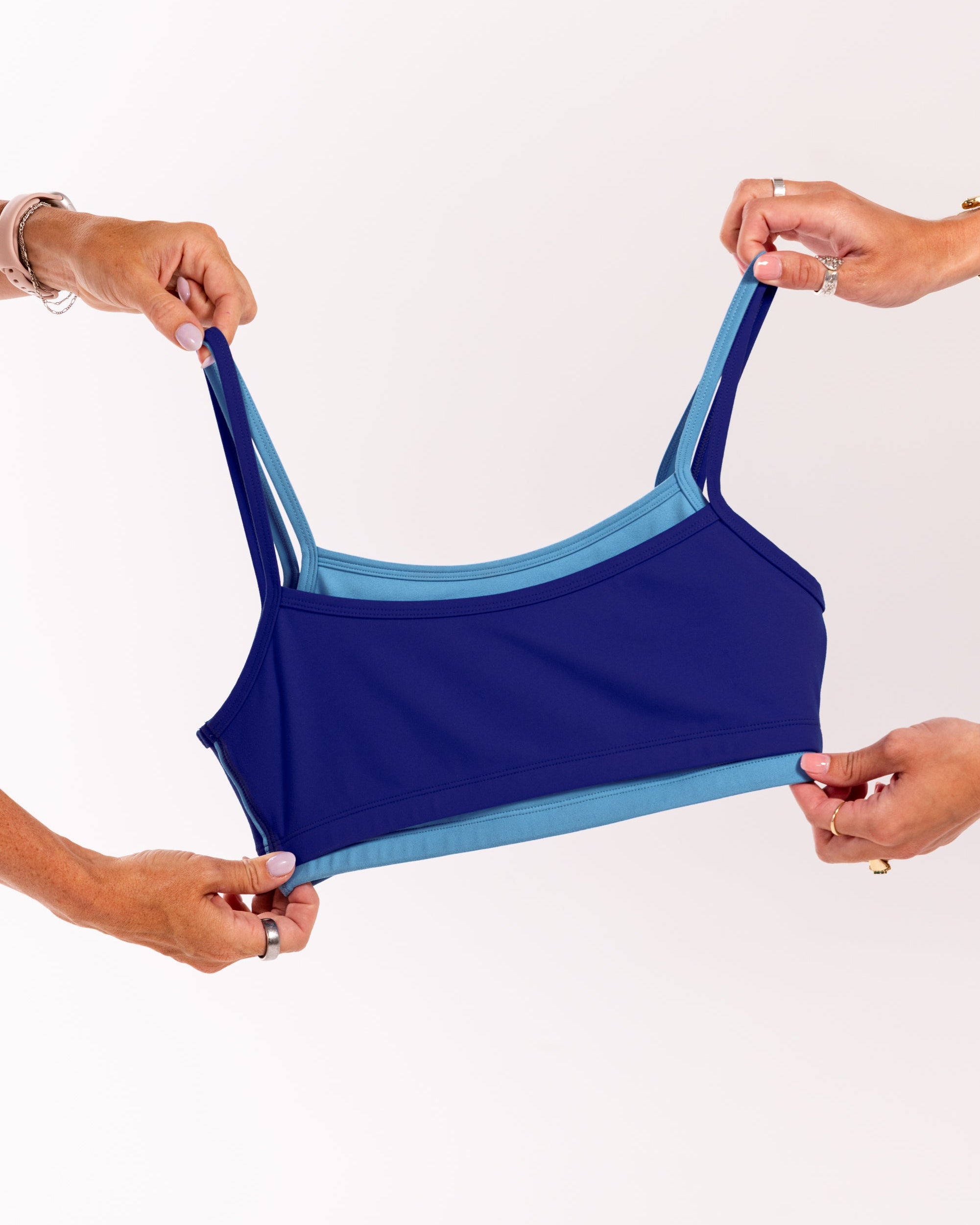 A navy blue sports bra with light blue trim and straps, held up by four hands against a plain white background.